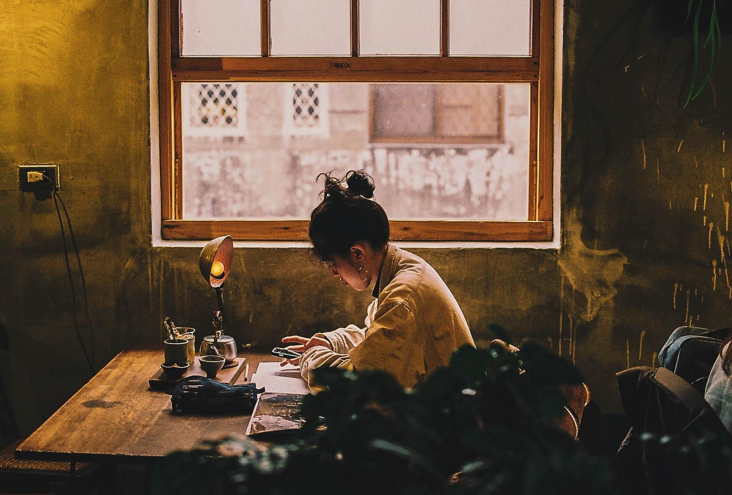 A woman sits at a wooden table with a lamp and tea set, looking at her phone next to a window.