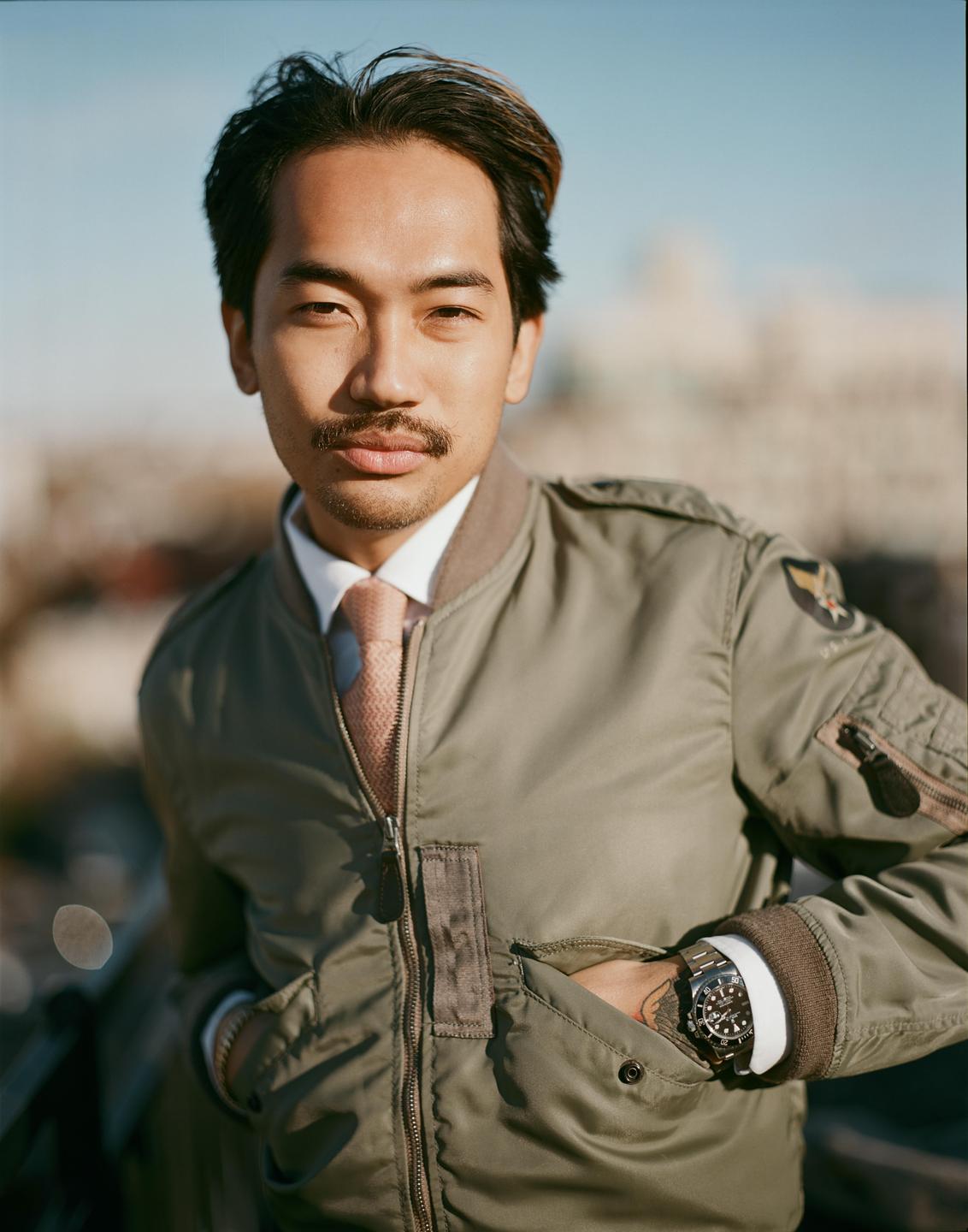 A man wearing a bomber jacket and tie looks directly at the viewer.