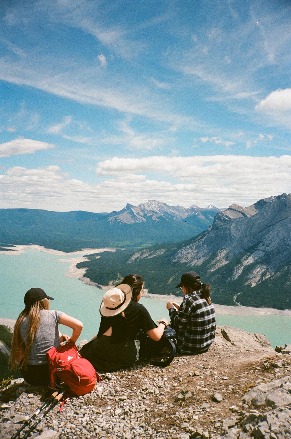 Three women sit on a rocky mountain peak overlooking a turquoise lake and distant mountains under a blue sky with clouds.