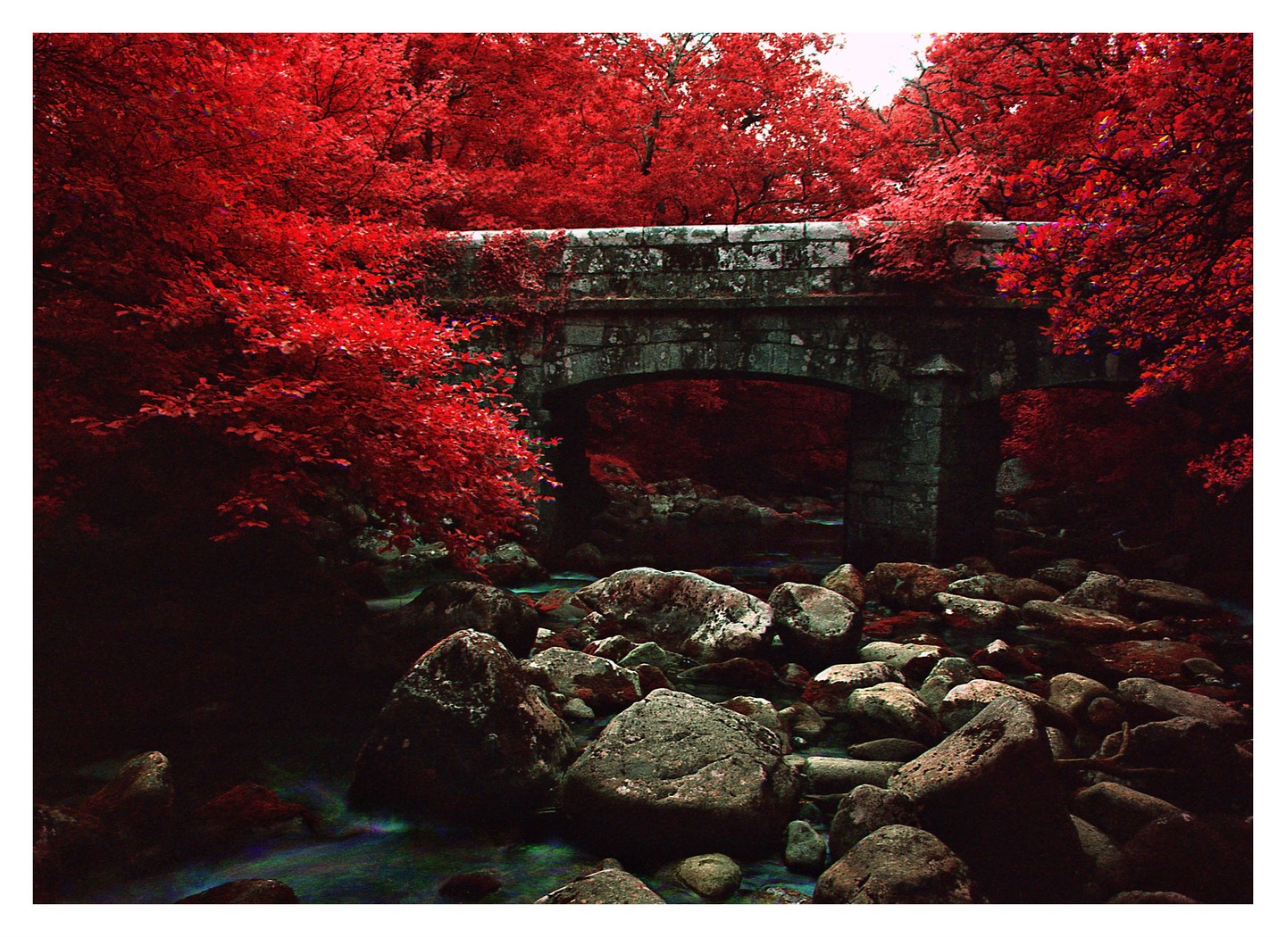 A stone bridge crosses a rocky river surrounded by trees with red foliage.