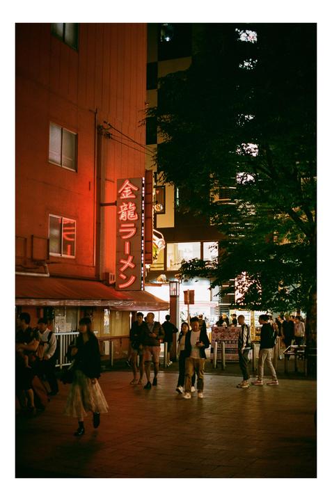 A street at night is filled with people walking past a building with a neon sign that reads "金龍ラーメン" (Kinryu Ramen).