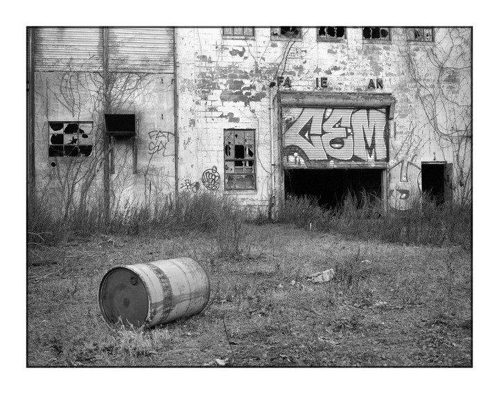 A barrel lies on its side in overgrown grass in front of an abandoned building with broken windows and graffiti on its roll-down door.