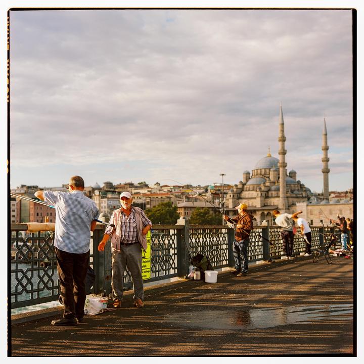 Men are fishing from a bridge with a mosque and city in the background.