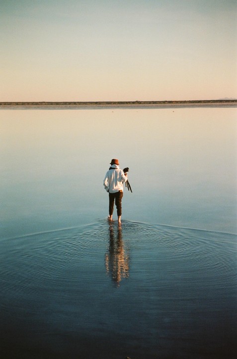 A person stands barefoot in shallow water holding a tripod, with a distant horizon line.