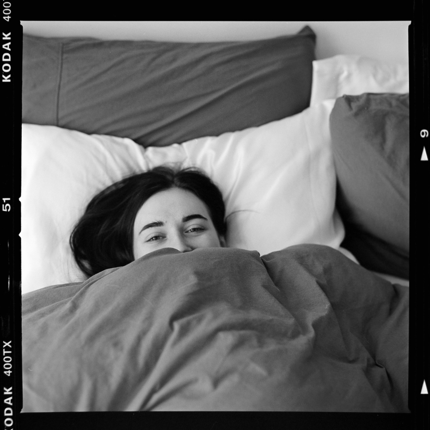 A woman lies in bed, peeking over a blanket.