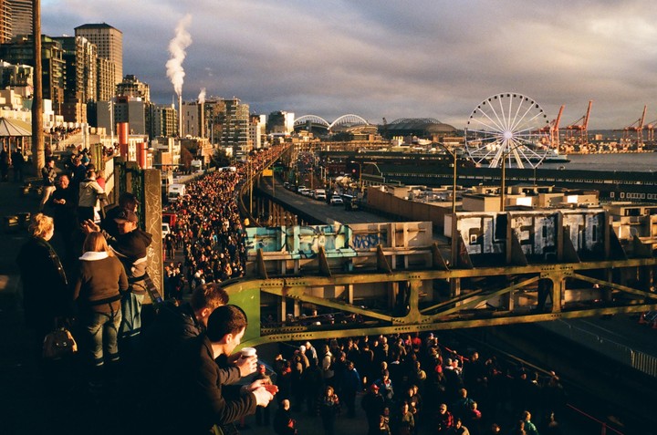 A large crowd of people gathers on a street overlooking a city with a ferris wheel and harbor.