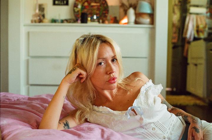 A woman with blonde hair and tattoos is lying on a pink bed.