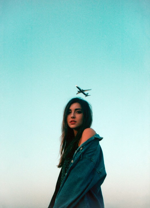A woman with long hair and a denim jacket looks at the camera with an airplane flying in the sky above her head.