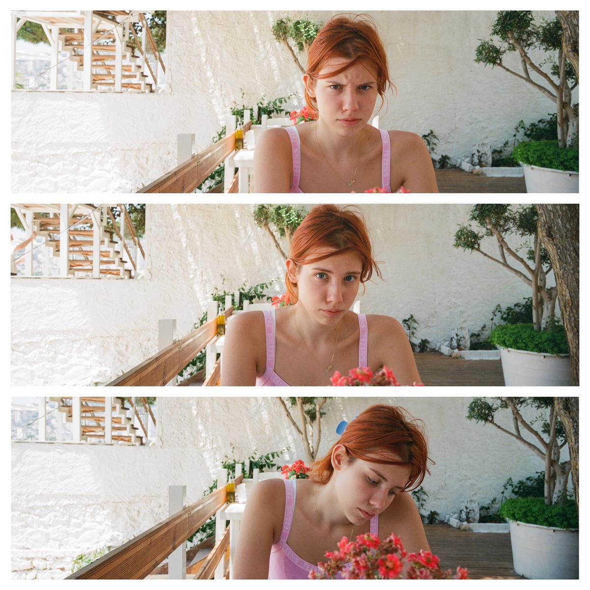 A woman with red hair sits at a table with red flowers in front of a white wall and a wooden railing.