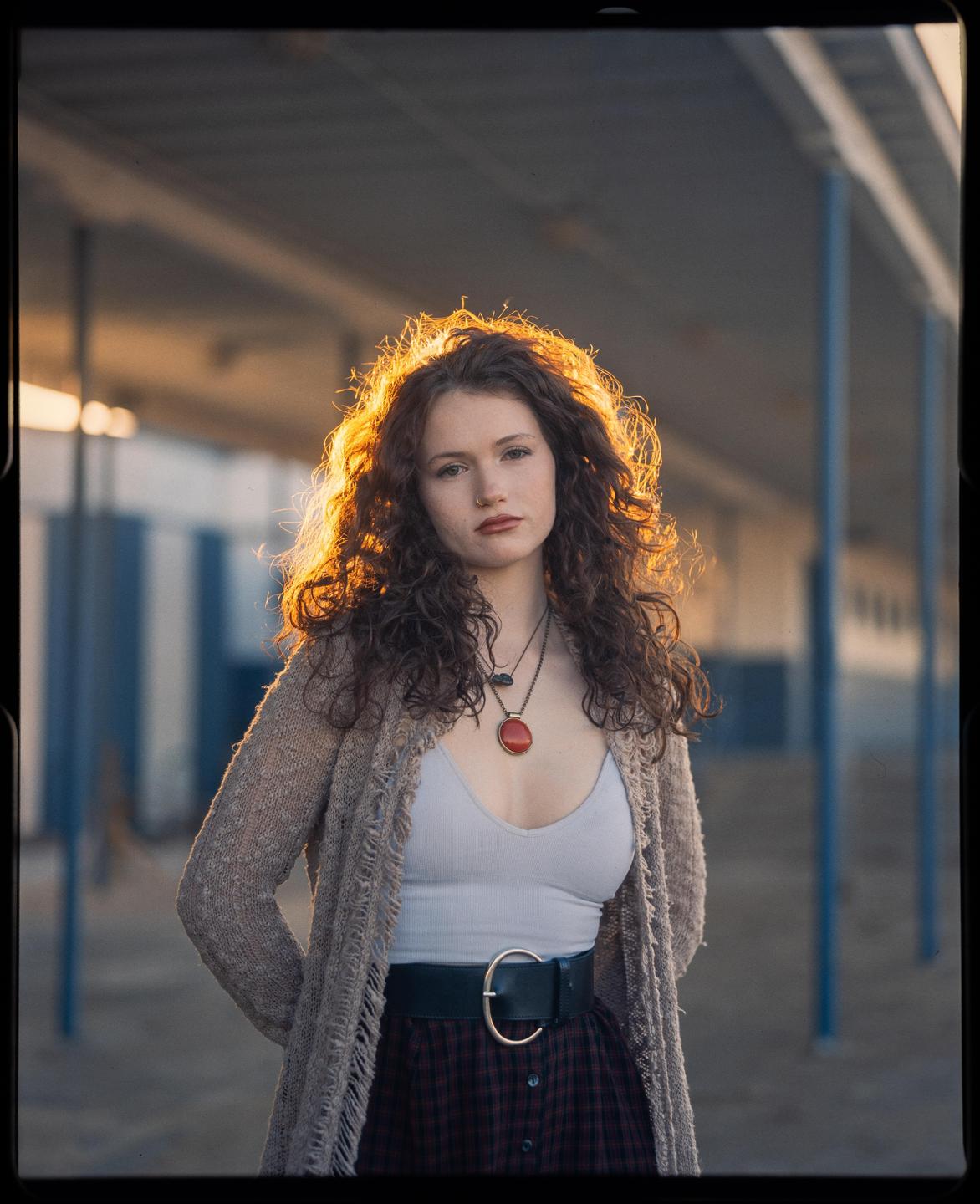 A woman with long curly hair stands backlit by the setting sun, wearing a cardigan, tank top, and plaid skirt.
