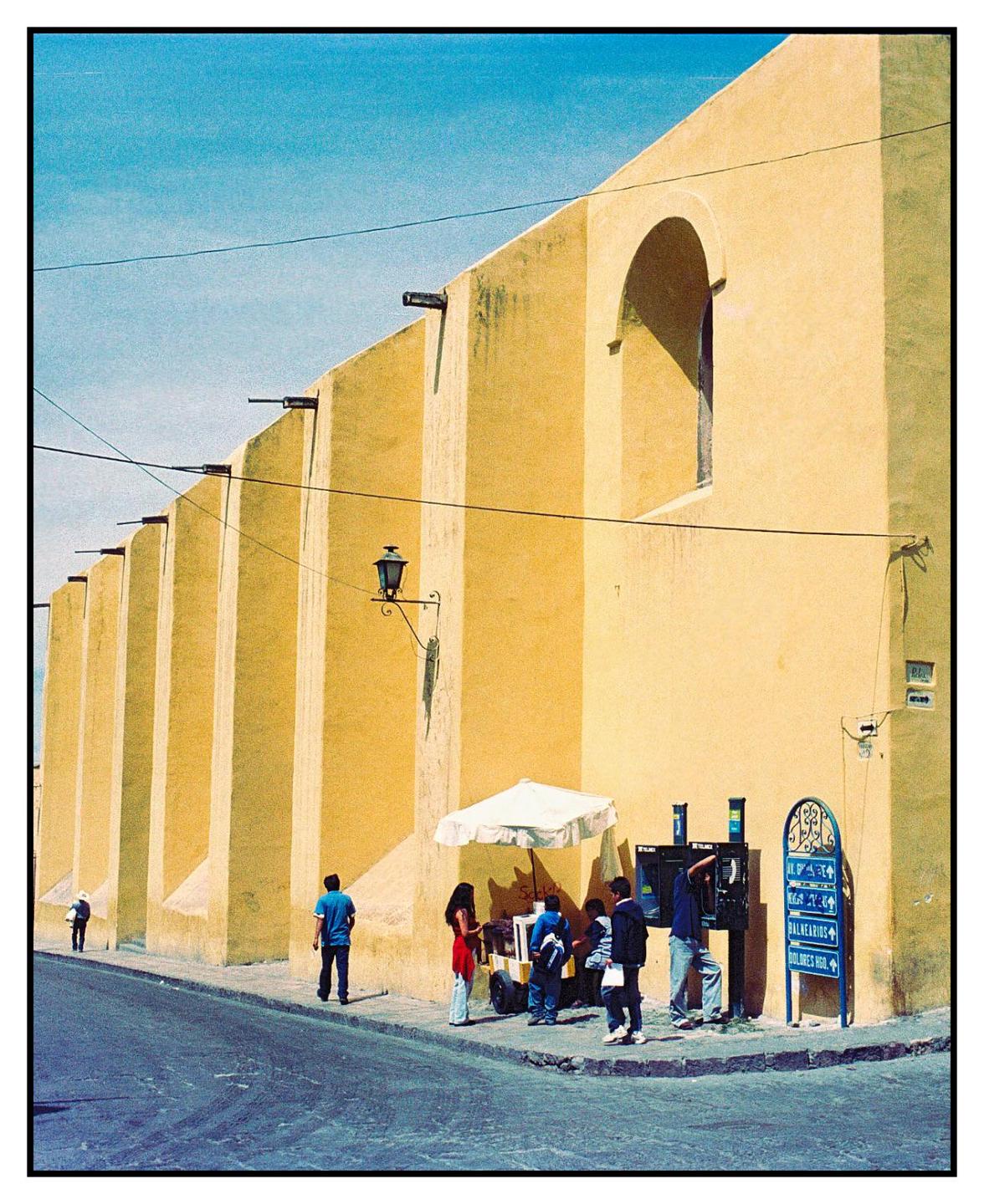 A group of people stand near a street vendor and public telephones next to a large yellow building.