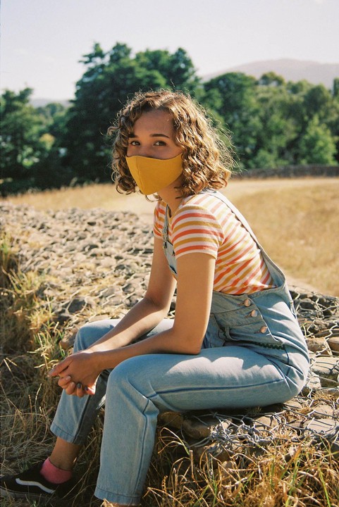 A woman wearing a yellow face mask and denim overalls sits on a stone wall in a field.