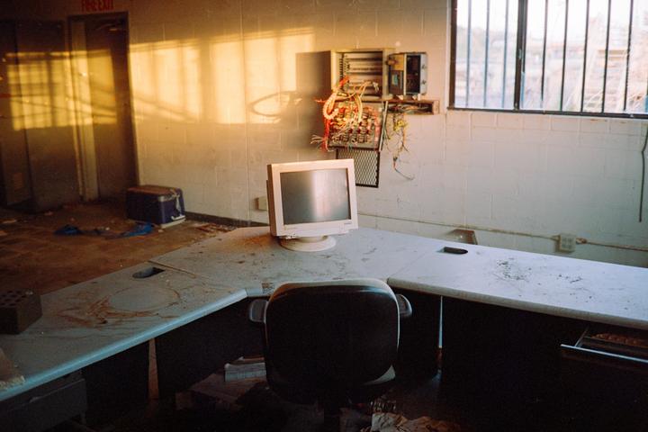 An abandoned office with a computer monitor on a dusty desk is illuminated by golden hour light streaming through a barred window.