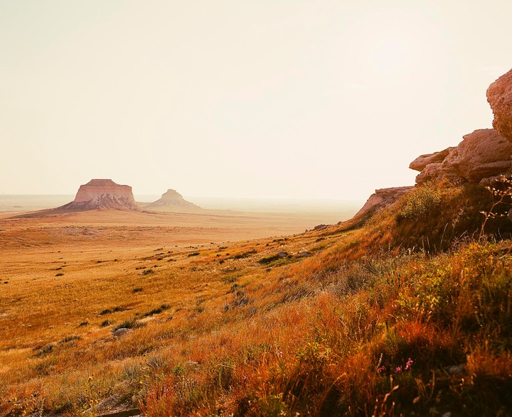 A vast, dry landscape with two buttes in the distance and a rocky, grassy slope in the foreground.