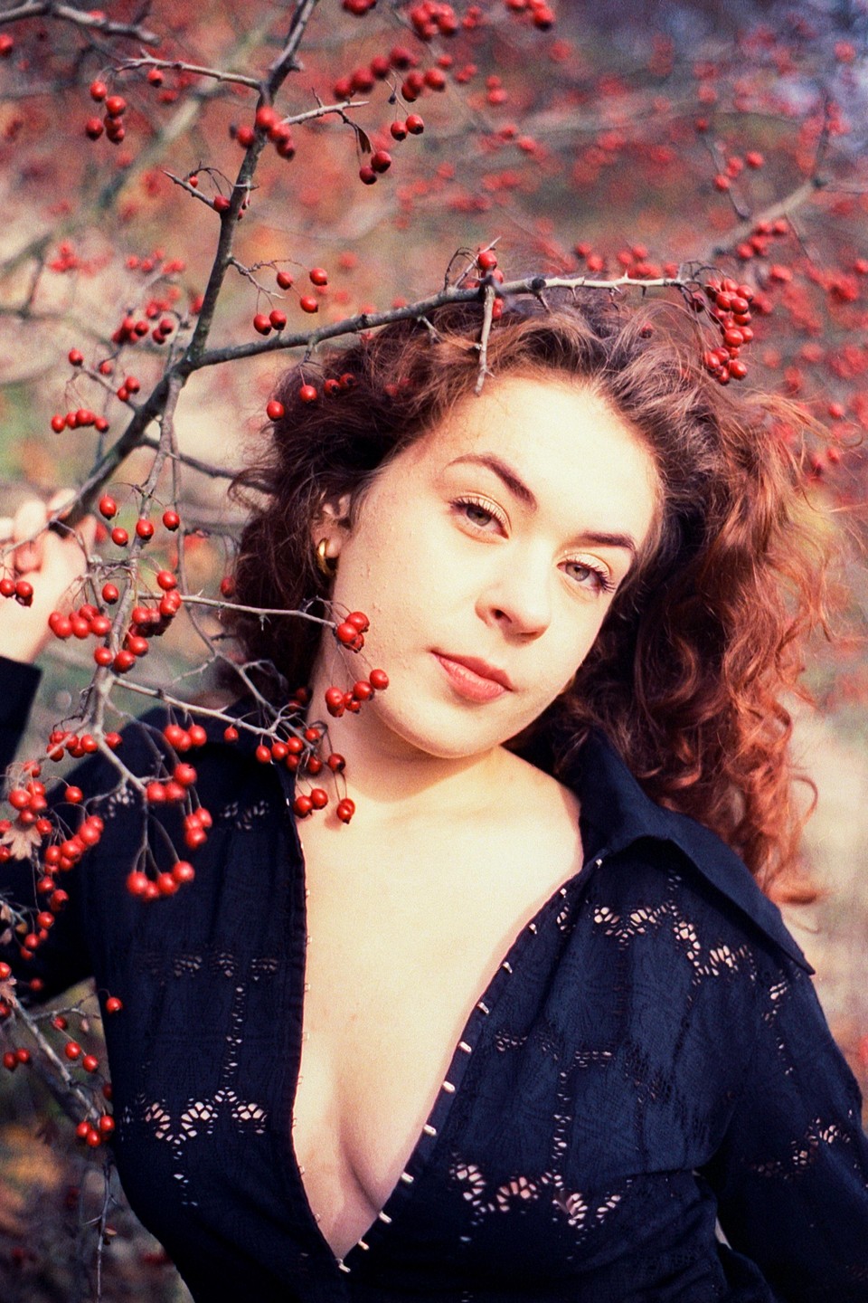 A woman with curly hair and a dark top stands among branches with red berries.