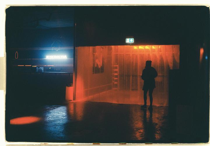 A person stands in a dark room with a neon sign and orange light.