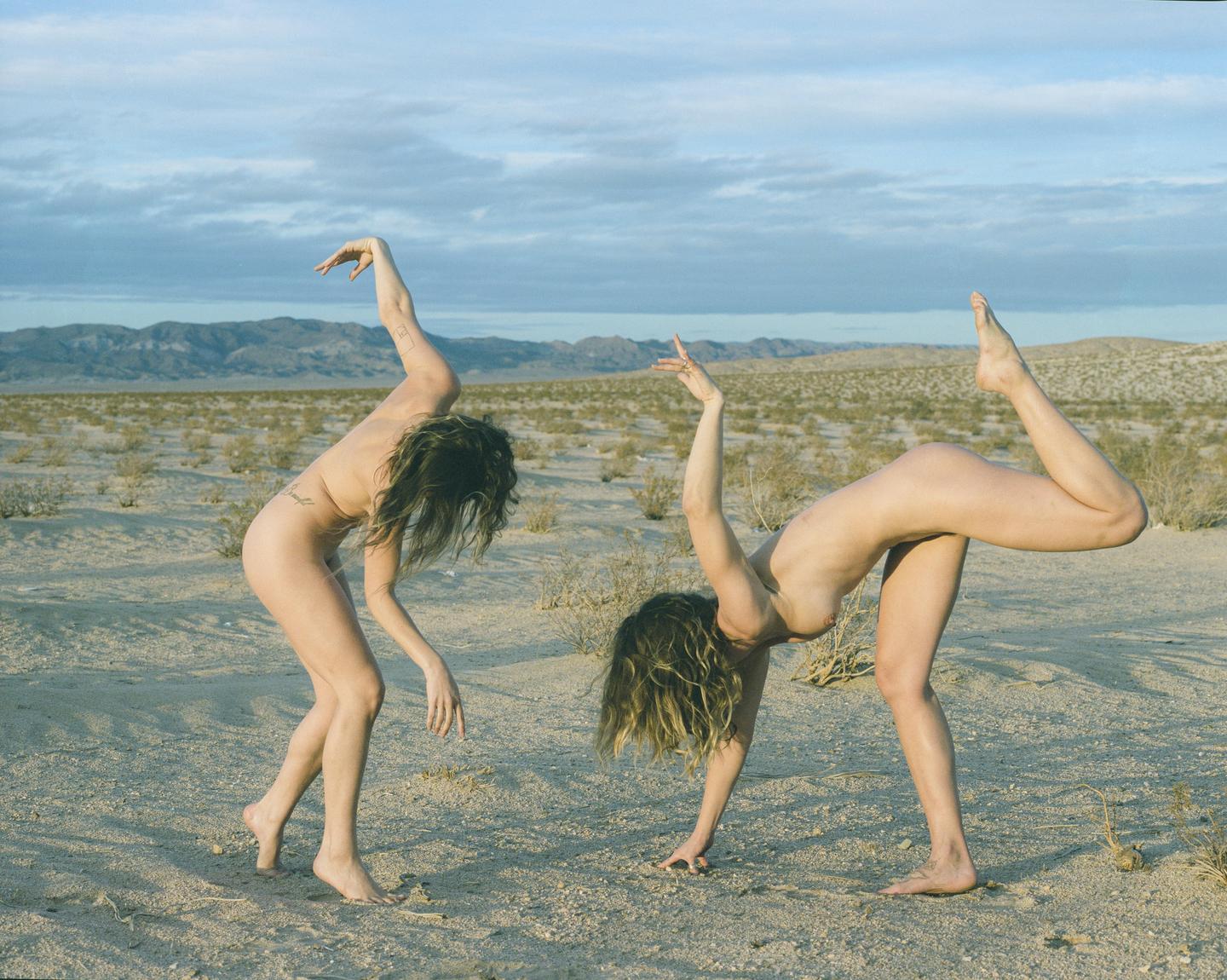 Two women pose nude in a desert landscape with mountains in the distance.