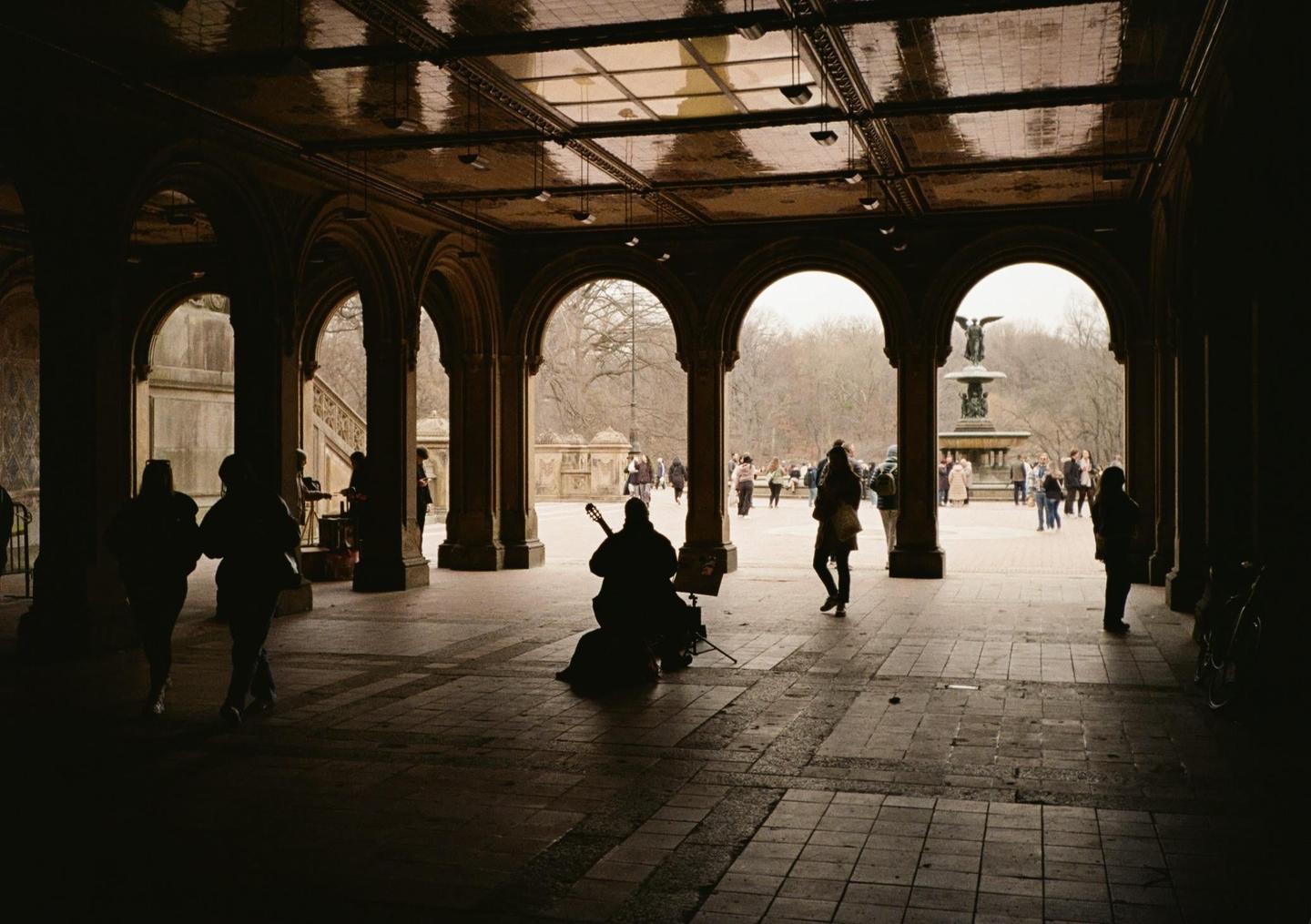 A musician plays guitar under an arched walkway with people and a fountain visible in the background.