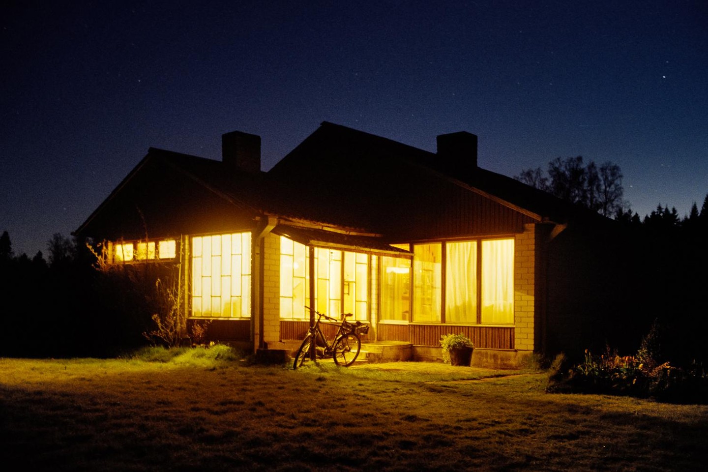 A house with illuminated windows and a bicycle parked outside is visible at night under a starry sky.