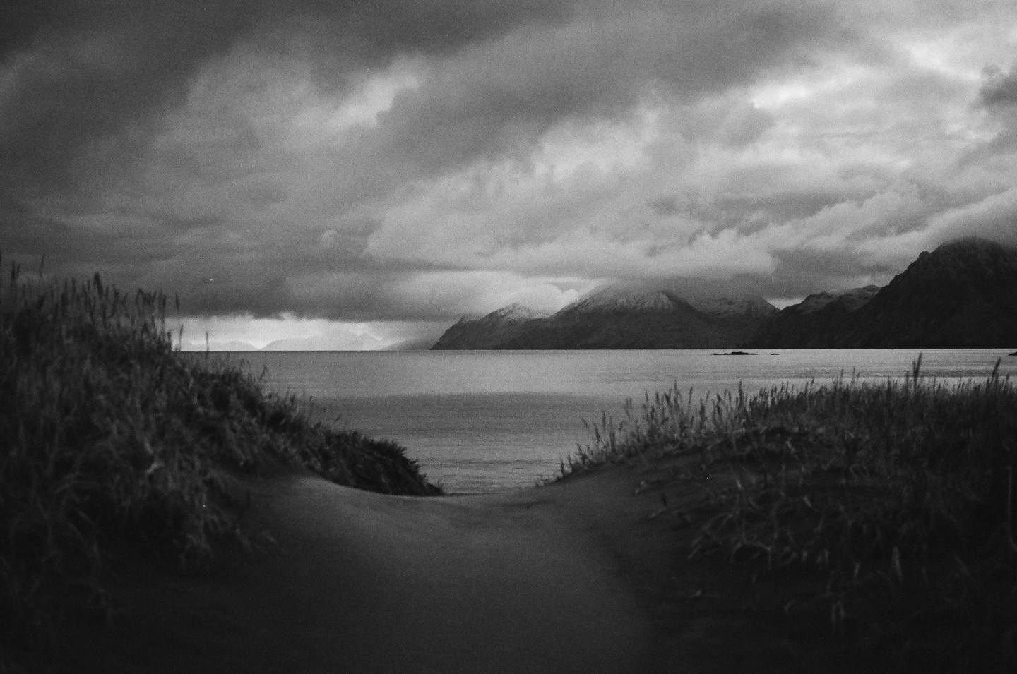 A path through grassy dunes leads to a body of water with mountains under a cloudy sky.