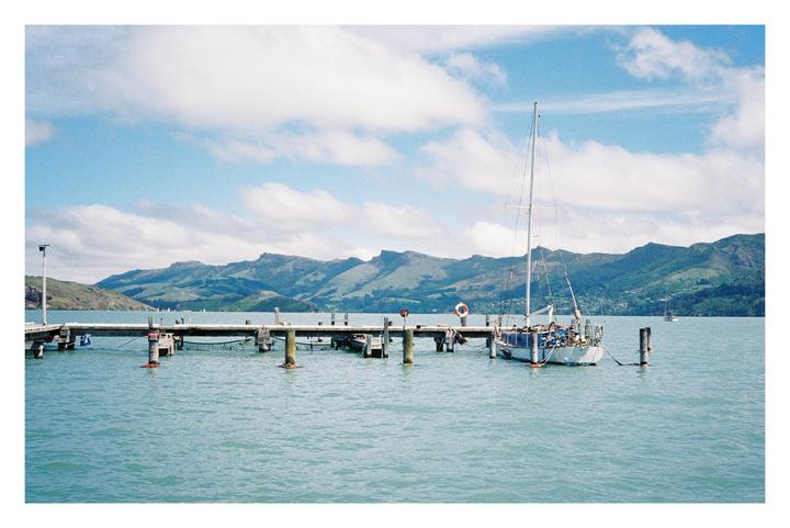 A sailboat is docked next to a wooden pier on a body of water with mountains in the background under a cloudy sky.