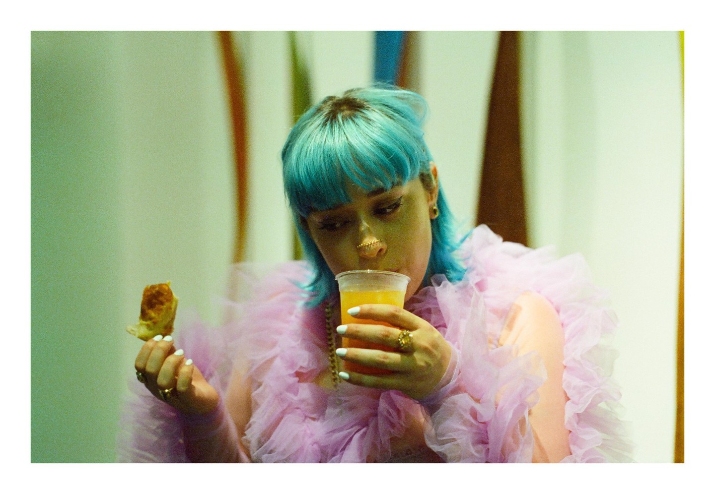A woman with blue hair and a ruffled pink top drinks from a plastic cup and holds a piece of food.