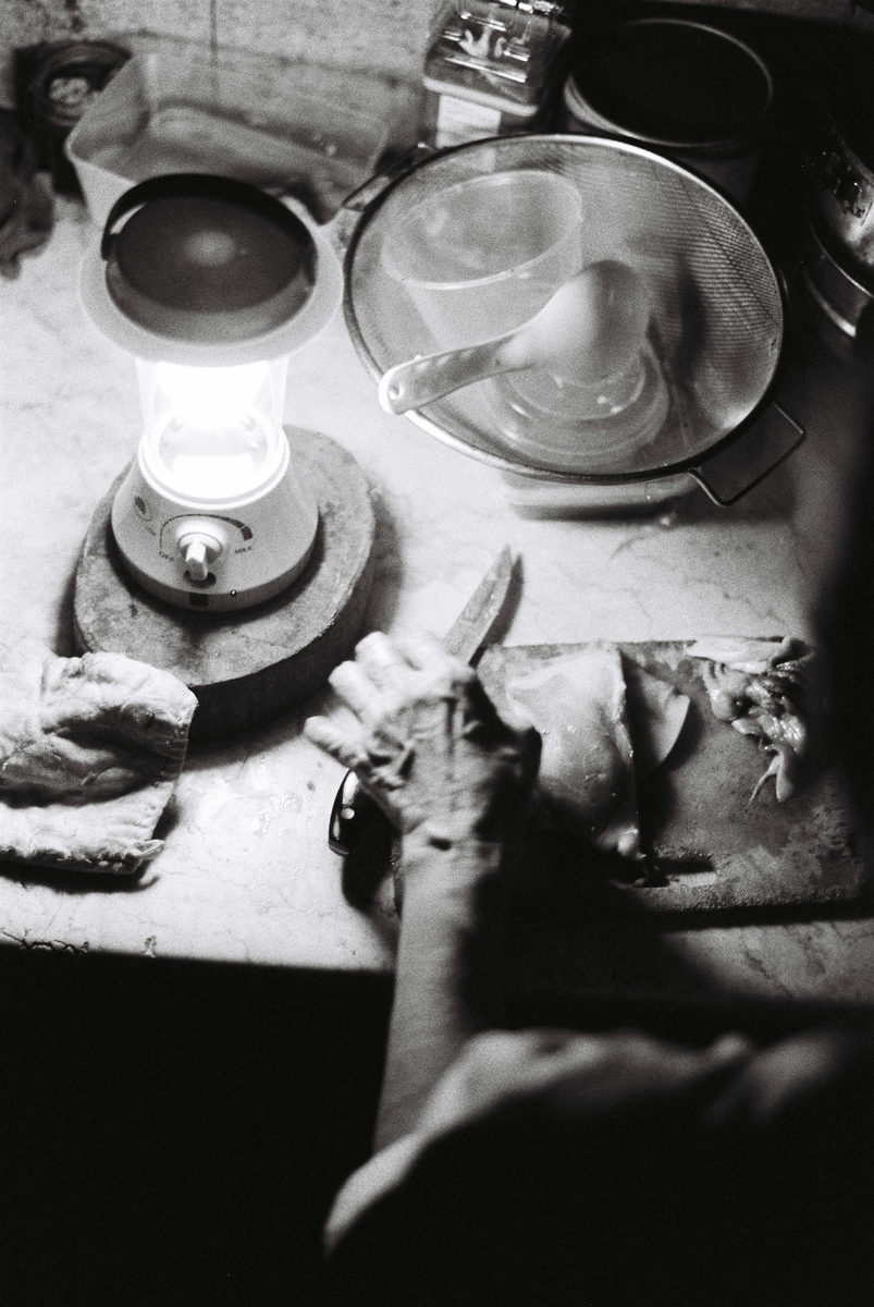 A person's hands are visible cutting meat on a wooden board next to a lit lantern and kitchen utensils.