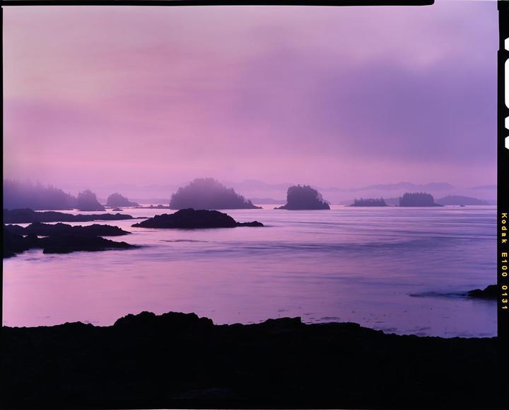 A purple sunrise illuminates a rocky coastline with small islands in the distance.