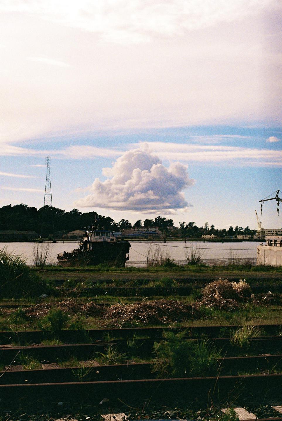 A boat is docked on a riverbank with overgrown train tracks in the foreground and a large cloud in the sky.