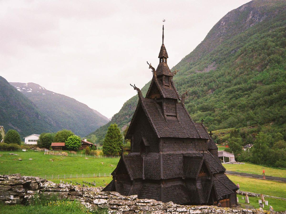 A dark wooden stave church with a tall spire stands in a green valley surrounded by mountains.