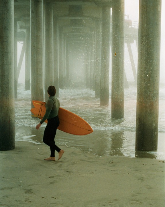 A surfer in a wetsuit carries a surfboard under a pier on a hazy day.