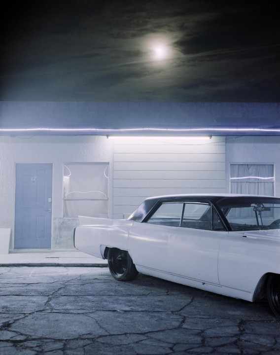 A white car is parked in front of a building with a blue door under a cloudy night sky with a visible moon.