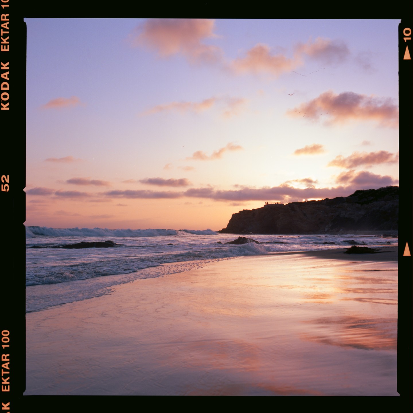 Waves crash on a beach at sunset, with a cliff and two people visible in the distance.
