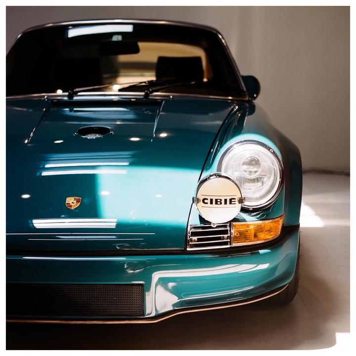 The front of a teal vintage car with a round headlight and a CIBIE auxiliary light is shown.