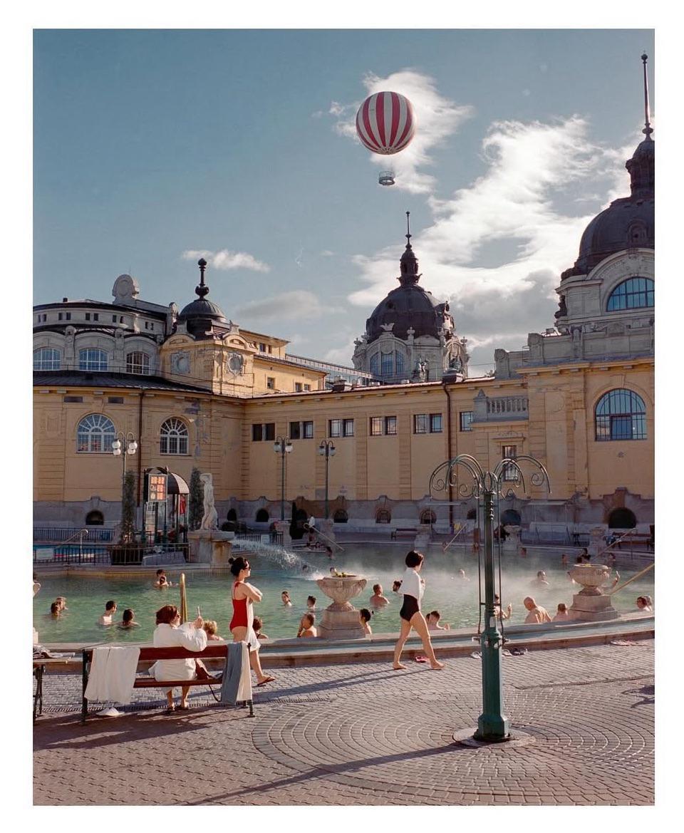 A hot air balloon floats above a thermal bath with people swimming and relaxing.