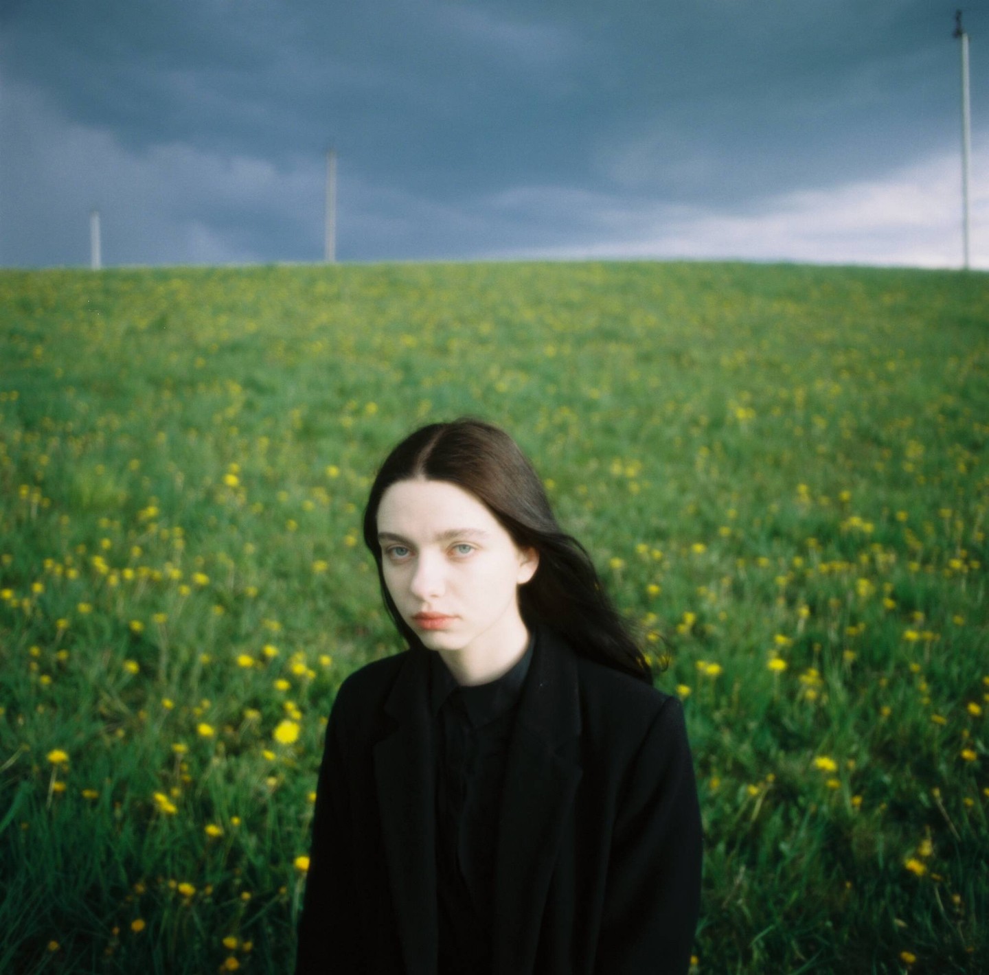 A woman with long dark hair stands in a field of green grass and yellow flowers under a dark, cloudy sky.
