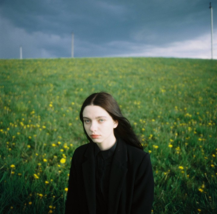 A woman with long dark hair stands in a field of green grass and yellow flowers under a dark, cloudy sky.
