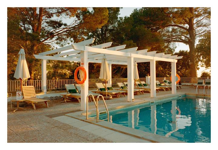 A swimming pool area with lounge chairs, umbrellas, and a white pergola is bathed in golden light.