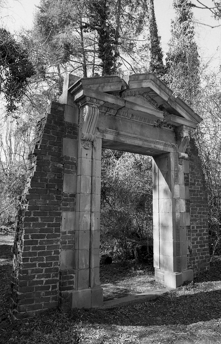 A stone archway with decorative carvings stands amidst overgrown foliage and trees.