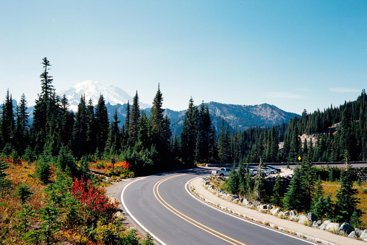 A winding road leads through a forest towards a snow-capped mountain under a clear sky.