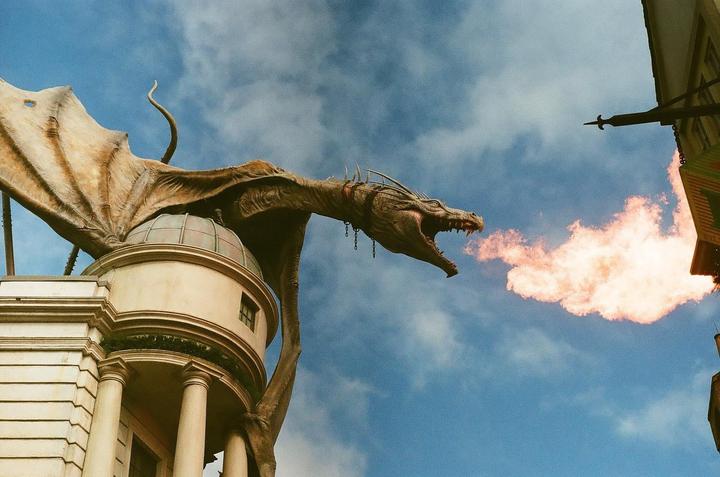 A large dragon statue with wings is perched on a building, breathing fire into a blue sky with clouds.