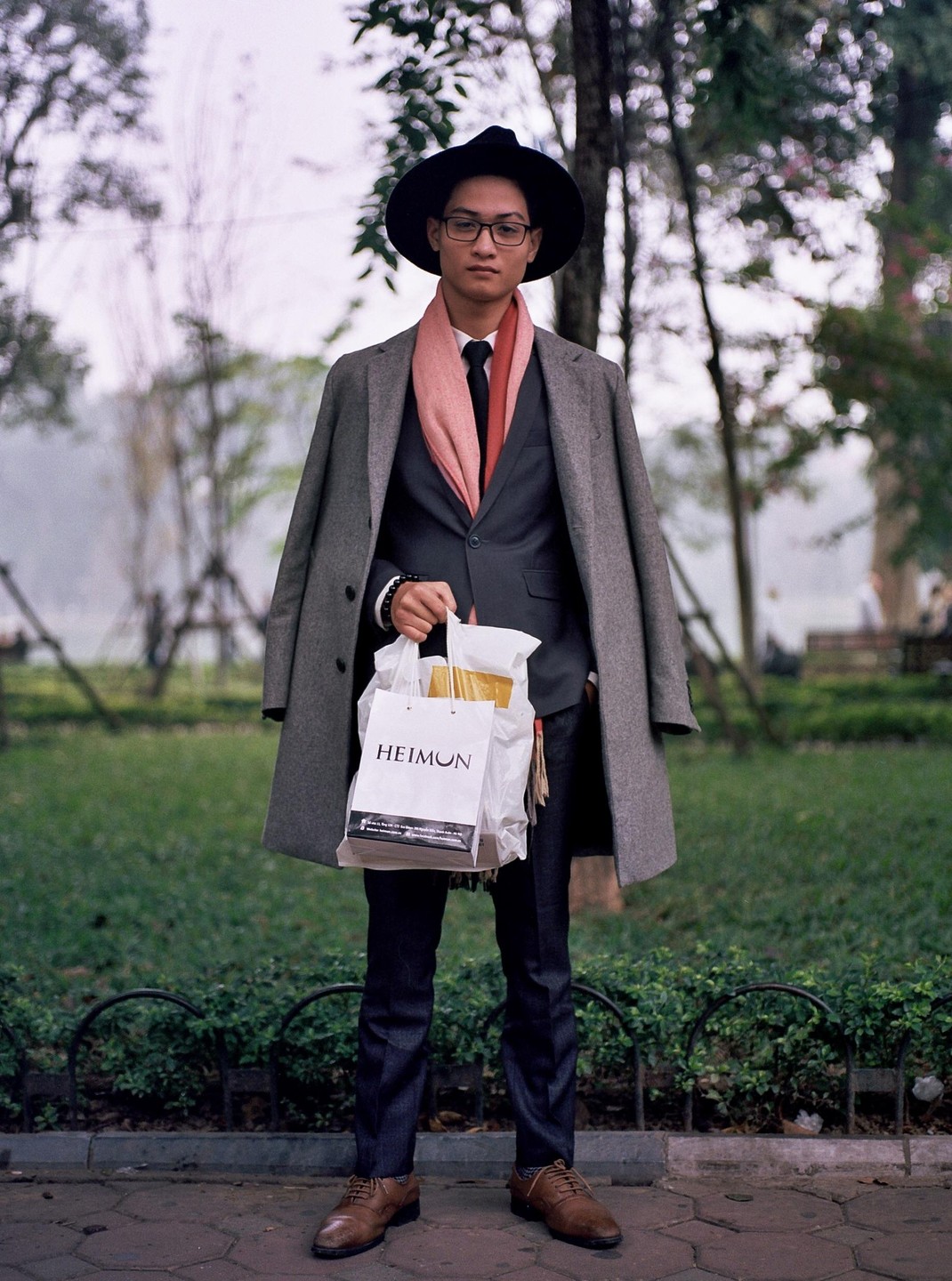 A man in a suit, coat, and hat stands on a sidewalk holding a shopping bag.