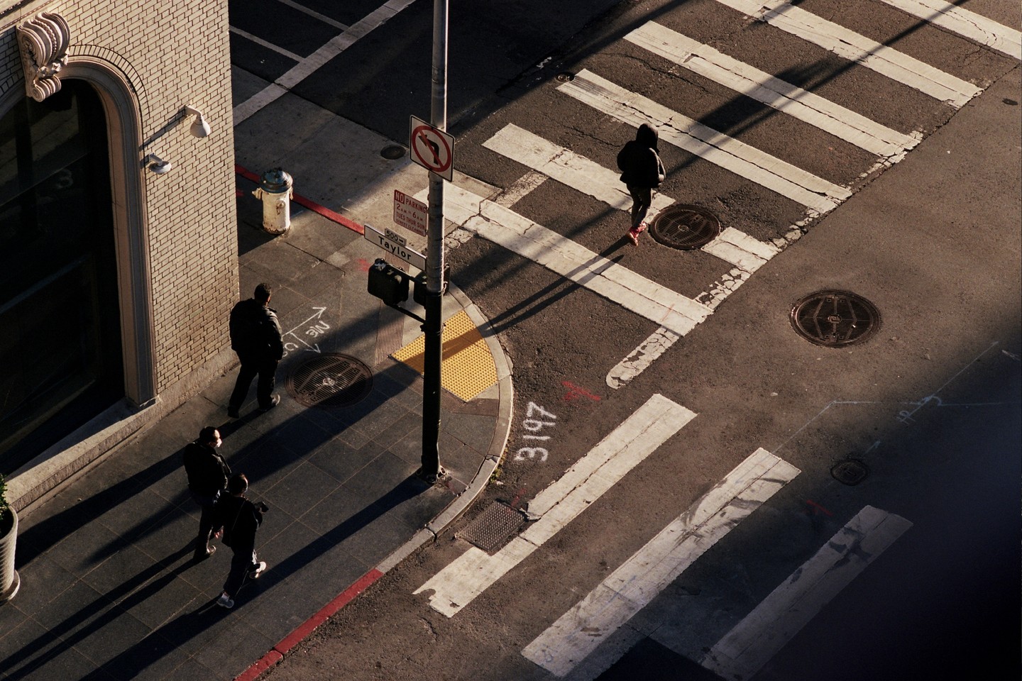 Three people walk on a sunlit street corner with crosswalks and a street sign.