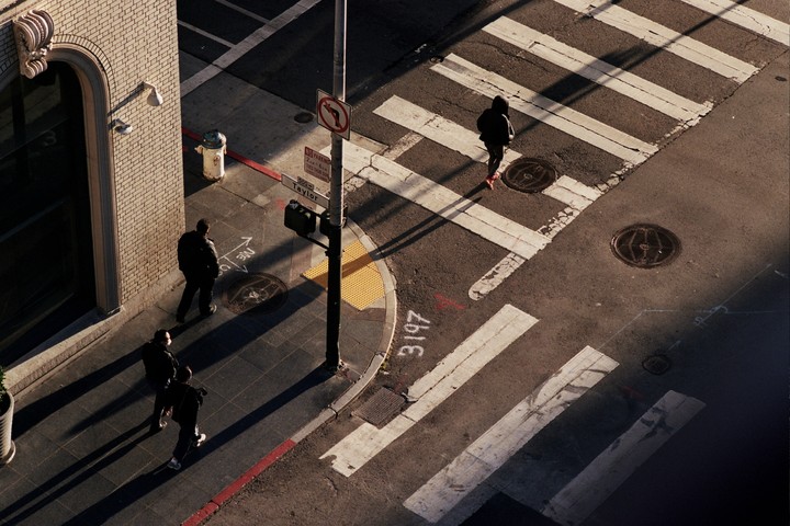Three people walk on a sunlit street corner with crosswalks and a street sign.