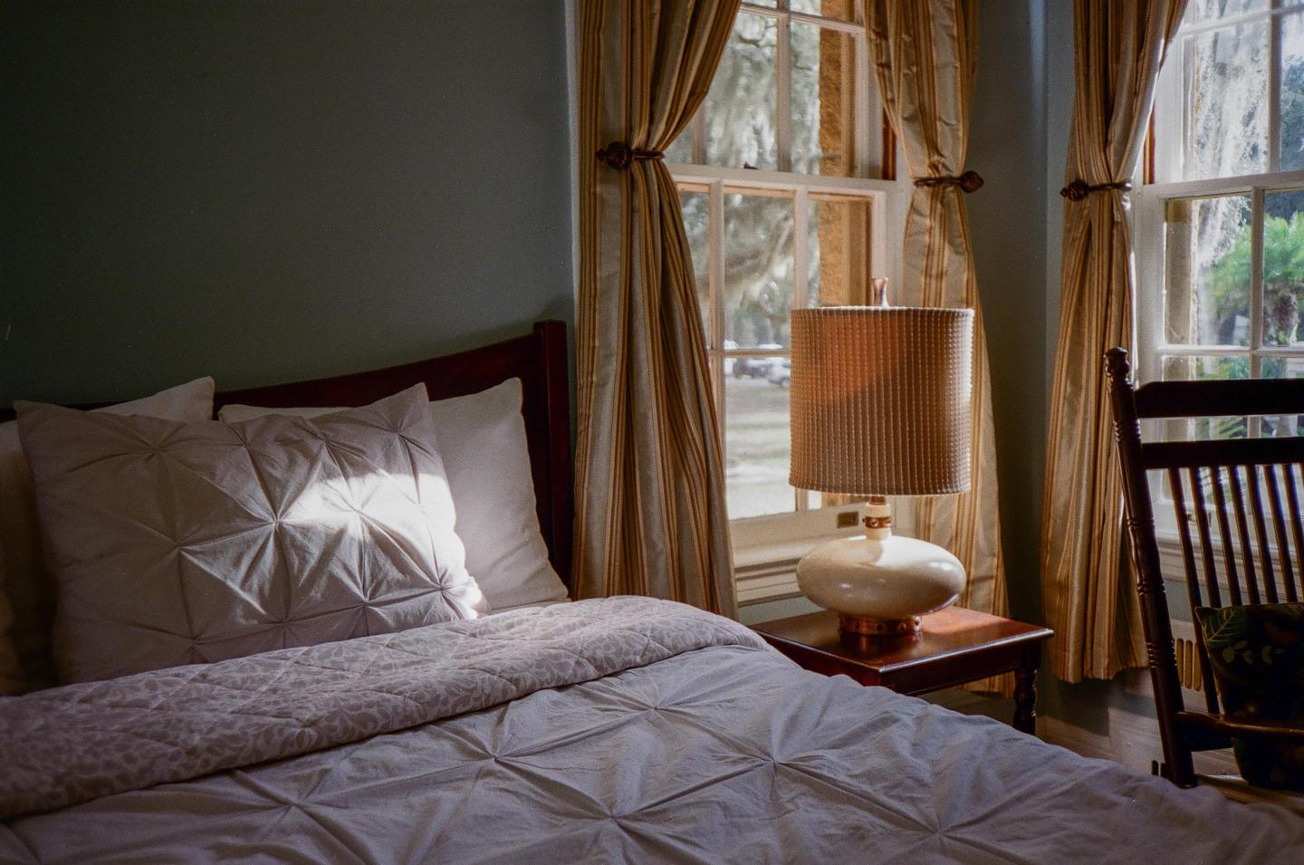 Sunlight streams through a window onto a bed with pillows and a patterned comforter, next to a bedside table with a lamp.