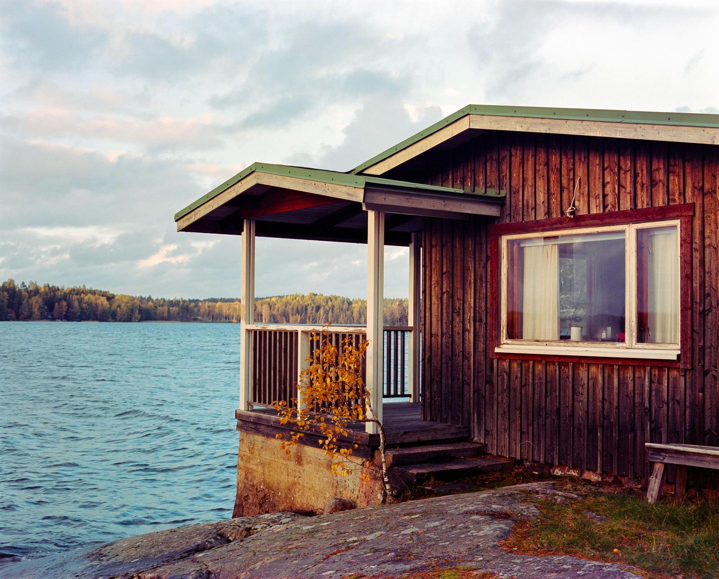 A wooden cabin with a porch sits on a rocky shore next to a lake with a forest in the distance under a cloudy sky.