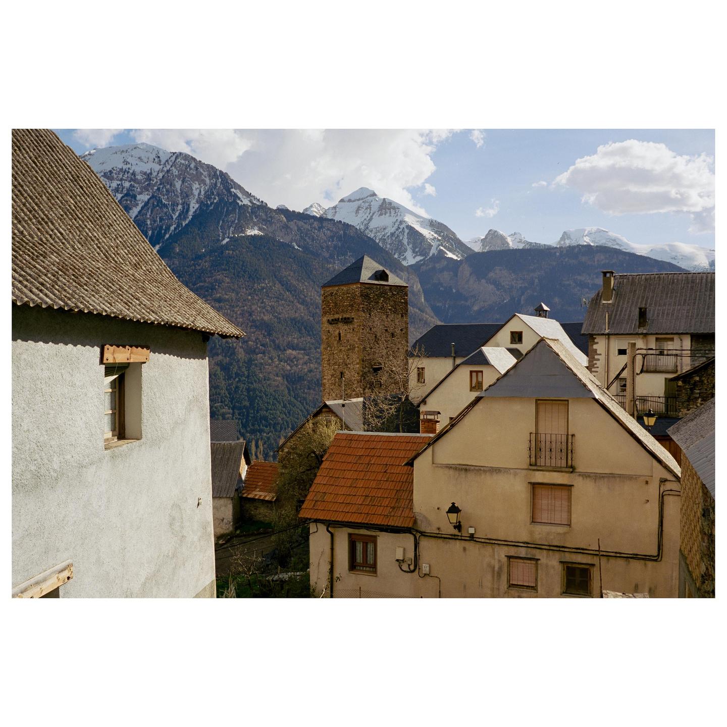 A village with a stone tower and houses with red and grey roofs is nestled in a valley with snow-capped mountains in the background.