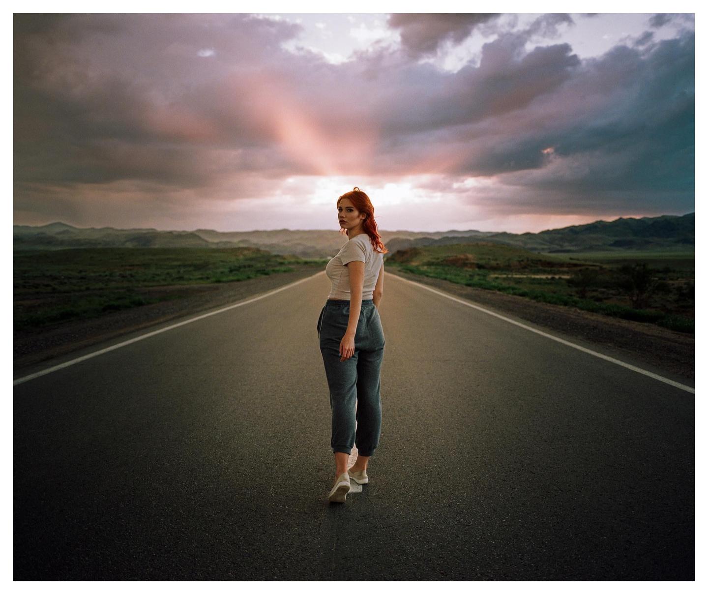 A woman stands on a long, empty road with mountains in the distance under a dramatic sky.