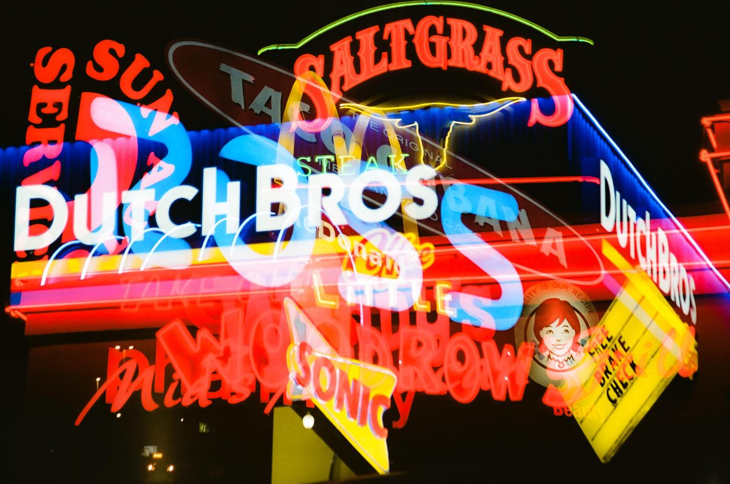 Multiple neon signs from various businesses are overlaid, creating a colorful and chaotic display against a dark background.