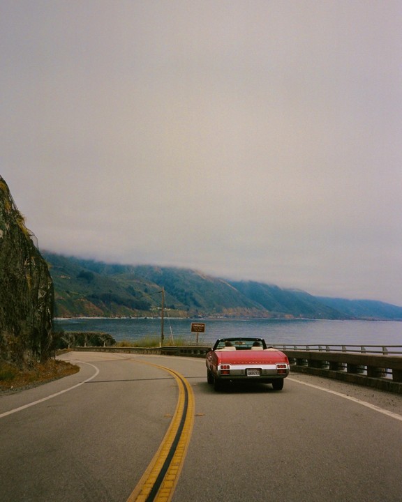 A red convertible drives along a winding coastal road with mountains and ocean in the background.